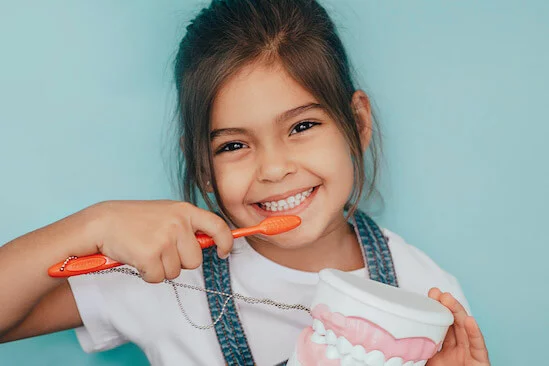 Child wearing white shirt and denim overalls, holding a dental model and demonstrating flossing with a dental floss tool. The dental model features artificial teeth used for educational purposes to teach oral hygiene.