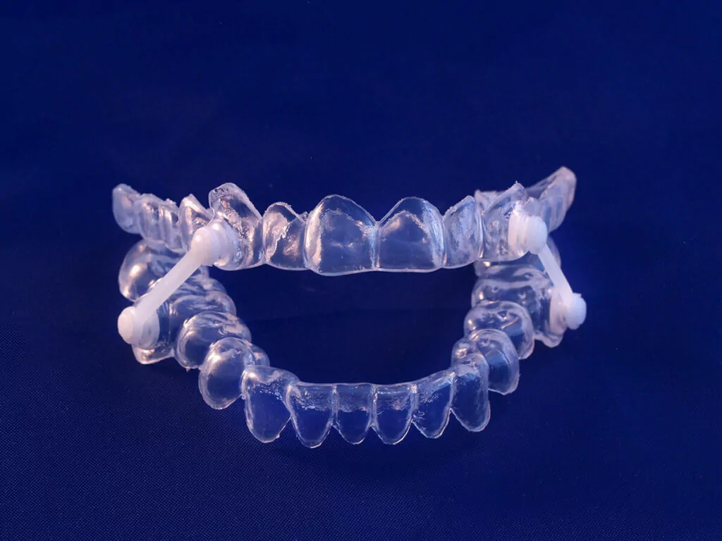 A pair of clear sleep apnea aligners with white attachments on a dark blue background.