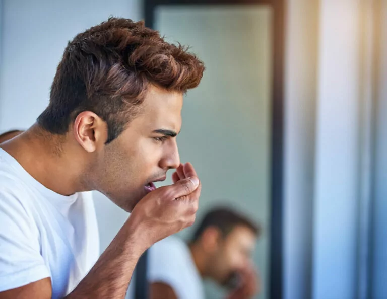 A person in a white shirt leaning forward and cupping a hand near their mouth to smell their breath, standing in front of a bathroom mirror with a blurred reflection in the background.