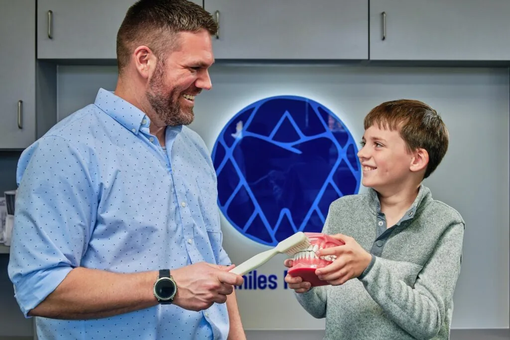 Two smiling individuals stand in a dental clinic, one holding an oversized toothbrush and the other displaying a model of teeth, as if demonstrating proper brushing technique.
