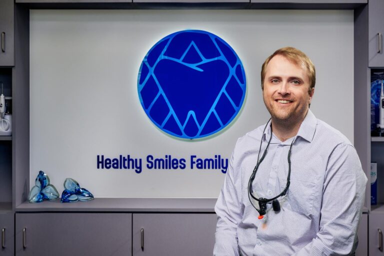 Dr. Joel Benson wearing a white shirt with a dental loupe sits in front of a “Healthy Smiles Family” logo featuring a tooth and heart icon inside a blue circle. Surrounding shelves with decorative glass objects and cabinets suggest a professional dental setting with branding elements subtly integrated into the backdrop.