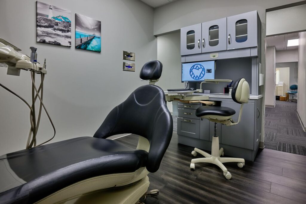 Modern dental examination room featuring a reclining black and beige dental chair, mounted dental tools, a swivel stool, cabinetry with a computer screen, and artwork on the walls. The space has clean, contemporary decor with dark wood flooring and grey walls.