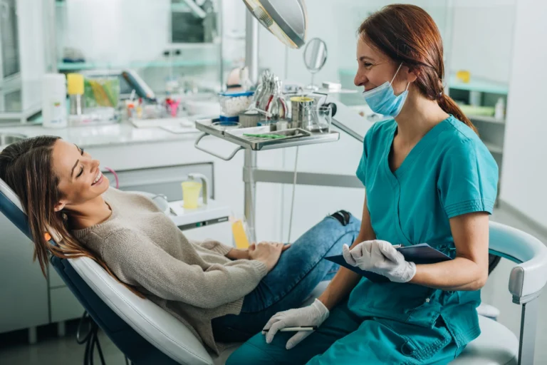 A patient in a beige sweater reclines in a chair while a dental professional in teal scrubs, gloves, and a face mask holds a clipboard giving out tooth extraction after care tips
